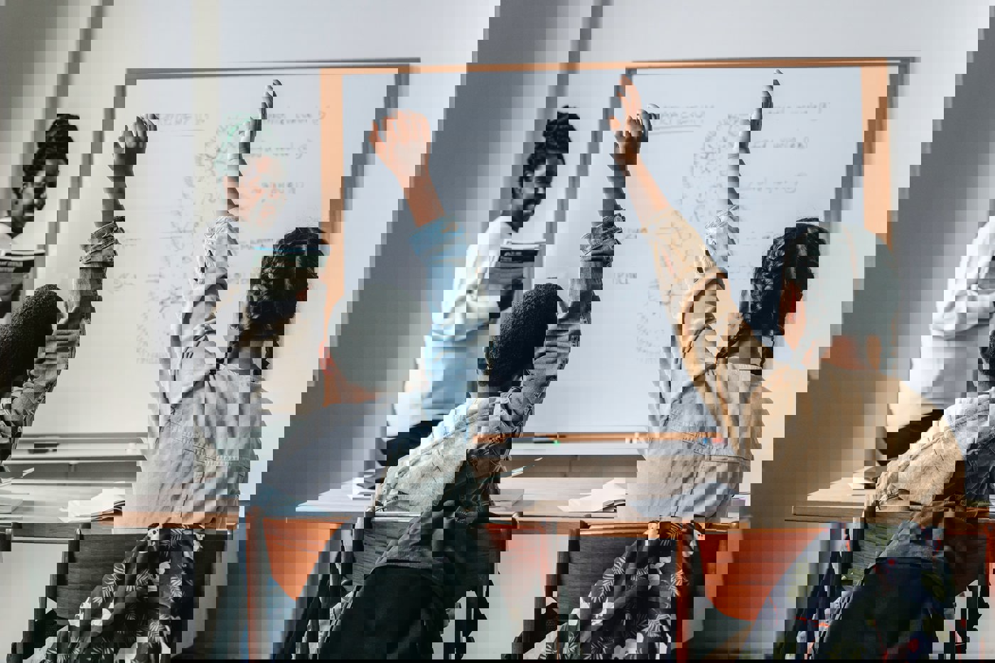 Image of two children in a school setting with their hands raised in front of a teacher and whiteboard